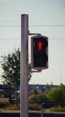 Bir direğe monte edilmiş yaya trafik ışığı kırmızı bir figür göstererek yayaların geçmemesi gerektiğini belirtirken, arka planda ağaçların ve geçen bisikletçilerin kısmi manzaraları yer alıyor.