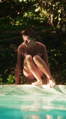 Renkli mayo giyen bir kız havuz kenarında zarif bir şekilde oturur, yemyeşil bitkiler canlı bir fon sağlar.