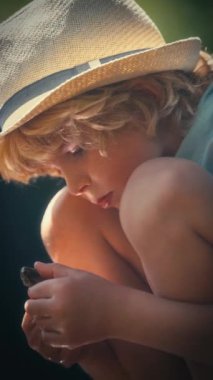 Güneş şapkası takan genç bir çocuk, yoğun bir merakla bir taşı inceler. Yüzü, yakındaki sudan yansıyan güneş ışığıyla aydınlanır.