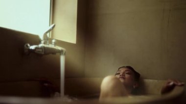 Bir kadın banyo küvetinde sırılsıklam olur, musluktan yavaşça akan su rahatlatıcı ve huzurlu bir atmosfer yaratır.