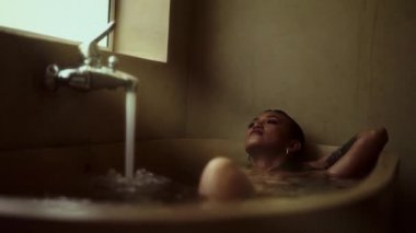 Bir kadın rahatlatıcı bir banyodan zevk alır, vücudunu usulca musluktan akan suyla küvette ıslatır.