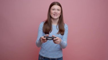 Mavi ve beyaz uzun kollu bluzlu bir kız, pembe bir duvara karşı gülümseyen ve eğlenen bir kumanda kullanıyor.
