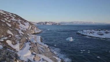 İnsansız hava aracı, karla kaplı kayalık adaları ve açık mavi gökyüzüne karşı okyanusta sürüklenen bereli parçaları gösteriyor.