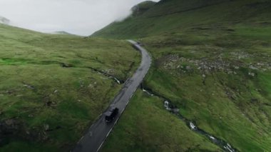 Sisli, engebeli yeşil bir arazide, uzak bir yolda ilerleyen siyah bir minibüs, aracın dramatik ve tenha bir arazide yolculuğunu vurguluyor.