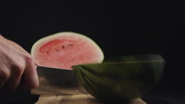 Keskin bir bıçak olgun bir karpuzu dilimliyor tahta bir tahtada, sulu kırmızı et ile siyah arka plan karşı karşıya