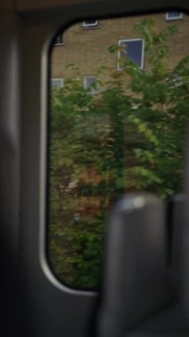 Hareket halindeki bir trenin penceresinden görüntülenen sahne bulanık bir yansımayı gösteriyor, dışarıdan yeşillik ve arka planda kısmen görünür tuğla bir bina ile hareket hissi ekliyor.