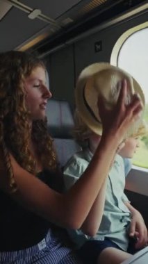 Kız kardeşi, tren yolculukları boyunca dikkatli bir şekilde kardeşinin güneş şapkasını düzeltiyor, hassas ve özenli bir an sergiliyor.
