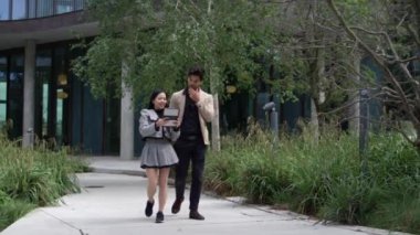 İki meslektaş, elinde tablet olan şık bir etekle dışarıda yürürken yanındaki adam da onunla sohbet ediyor.