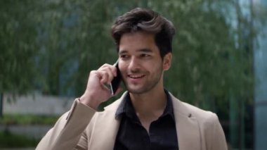 Koyu renk, hafif dalgalı saçlı, siyah bir gömleğin üzerine bej ceket giymiş, açık havada yeşil bir ortamda akıllı bir telefonla konuşurken gülümseyen bir adam.