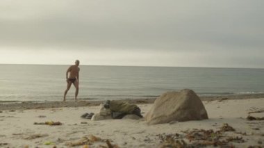 Sahilde yürüyen bir adam arka planda deniz olan plaj kıyafetlerine doğru yürür.