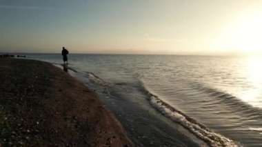 Gündoğumunda kıyıya vuran dalgalarla, sakin bir deniz kıyısında yürüyen bir adam.