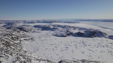 Kar tarlalarındaki karlı dağların ve kayalık çıkıntıların görüntüsü, açık mavi bir gökyüzü altında geniş araziyi ve engebeli özellikleri yakalıyor.