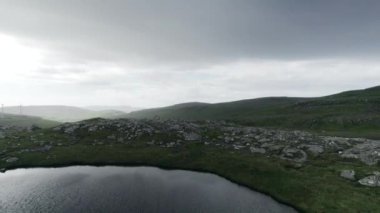 Bir nehir engebeli bir dağ manzarası boyunca akar ufukta uzak rüzgar türbinleri, doğal ve modern elementleri harmanlar.