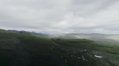 Sisli gökyüzünün altında yuvarlanan yeşil tepeler, sakin ve atmosferik bir manzara yaratır.
