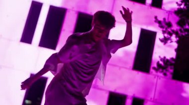 Genç bir erkek dansçının, pembe ışıklı ön cephe önünde hip hop hareketleri yaparken, dinamik enerjiyi ve sahnenin canlı ışıklarını yakaladığı Hollanda açılı bir çekim.