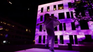 Genç bir adam canlı pembe ışıkla aydınlatılmış bir binanın ön cephesinde hareketli bir hip hop dansı yapıyor. Hareketlerini vurguluyor ve çarpıcı bir görsel karşıtlık oluşturuyor.