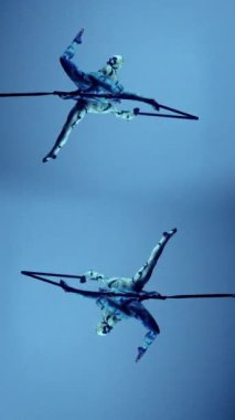 Akrobatlar etkileyici bir performans sergiliyorlar, sert bir çerçeveye bağlı halkalar ile sırt sırta asılılar, vücutları yatay olarak bir ayna görüntüsünde uzanıyor, bacakları V şeklinde ikiye ayrılıyor.