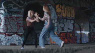 İki kadın ağır duvar yazısıyla kaplı bir arka fona saldırarak çarpışıyor.