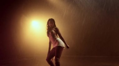 Arka planda sıcak turuncu ışıklarla aydınlatılmış, yağmurda dans eden bir kadın büyüleyici ve duygusal olarak yüklü bir atmosfer yaratıyor.