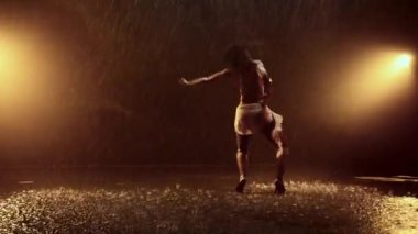 Enerjik bir kadın, yoğun yağmurda serbest stil dansı yapıyor. Arka planda parlak turuncu ışıklarla çarpıcı bir şekilde aydınlatılıyor ve büyüleyici ve dinamik bir sahne yaratıyor.