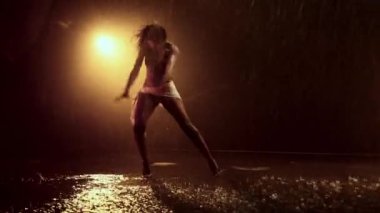 Yetenekli bir bayan dansçı etkileyici hareketlerini, parlak turuncu ışıklarla aydınlatılmış, yağmurla ıslanmış çarpıcı bir performansla sergiliyor.