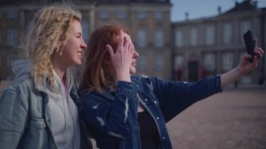 İki arkadaş yan yana durur, Amalienborg Sarayı 'nın görkemli arka planında selfie çekerken gülümserler.