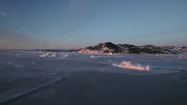 Uzaktaki karlı bir dağa yaklaşan bir dron, çevresindeki buzdağları ve deniz buzlarını çerçeveliyor.