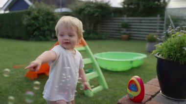 Genç, sarışın bir çocuk, kumsalın yakınındaki çimenli bir bahçede, sade beyaz bir gömlek giymiş ve plastik bir oyuncaktan neşeyle köpük kovalıyor.