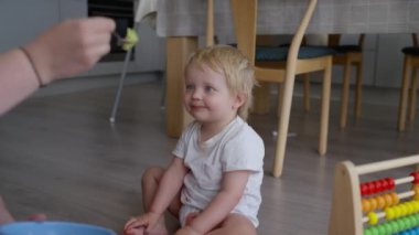 Evdeki mutfağın zemininde oturan tahta sandalyeli ve arka planda bir masa olan bir çocuk, bir yetişkinin neşeyle tuttuğu kaşıktan yemek yemek için öne doğru eğilir.