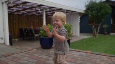 Çizgili bir gömlek ve pantolon giymiş neşeli bir sarışın çocuk, baloncuklar arasında oynarken heyecan verici bir şekilde el çırpıyor tuğla bir veranda, saksı bitkileri ile çevrili, otlak bir alan