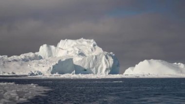 Kuzey Kutbu 'nun sakin ve dondurucu suları tarafından çerçevelenen uzak, devasa buz oluşumlarından kademeli olarak uzaklaşmayı belgeleyen el kameralı bir çekim.