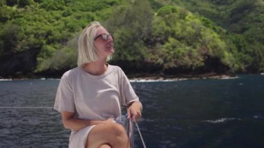 Kısa sarı saçlı, güneş gözlüklü ve açık renk gömlekli bir kadın, bir teknenin kenarına oturmuş, sakin bir yolculuğun tadını çıkarıyor, rahat duruşu ve nazik gülüşü.
