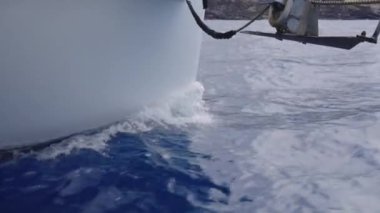 Okyanusta yüzen bir teknenin yakın çekim görüntüsü, pruvanın etrafında hafif dalgalanmalar oluşturan sakin su ve derin mavi denizle keskin zıt düşen beyaz gövdeye odaklanır.