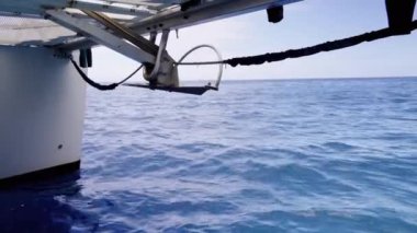 Bir zincire bağlı bir çapa, açık mavi okyanusa, gövdesi soldan görülebilen bir tekneden indirilirken, sakin su, hafif dalgalarla güneş ışığını yansıtıyor.