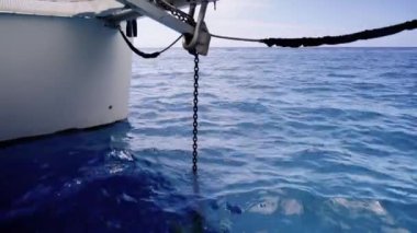 Bir zincire bağlı büyük, kıvrımlı bir çapa, açık mavi bir gökyüzünün altında, bir teknenin kenarındaki makara sistemi vasıtasıyla derin mavi okyanustan çekiliyor.