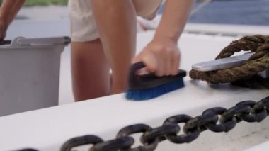 Yakın plan çekimde, mavi tüylü bir fırçayla güverteyi ovalayan bir kadının eli ön plana çıkarken, kısmen görünür bir kova ve ağır metal bir zincir tekneyle bağlantılı olduğunu gösteriyor.