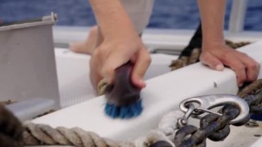 Yakından çekilen bir fotoğraf, arka planda bir halat, zincir ve metal kanca görünürken, mavi tüylü bir fırçayla güvertenin beyaz yüzeyini ovalayan bir kadının elini yakalar.