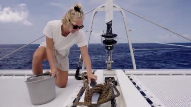 Beyaz tişörtlü ve açık renk şortlu bir kadın bir teknenin güvertesinde diz çöker, saçları topuz ve güneş gözlüğü içinde geri çekilir ve temizliğe odaklanırken gözlerini güneşten korur.