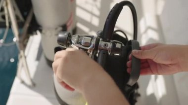 Yakın plan çekimde, güvenli bir şekilde bağlanmış, parlak metal bir scuba tüpü regülatörü ve hortumuyla, tank ve teknenin bulanık detaylarını gösteren, el ele tutuşur ve ayarlanır.