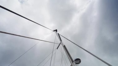 Yelkenlinin alçak açılı görüntüsü direği ve donanımı yakalar, bulutlu, bulutlu bir gökyüzüne uzanırken yüksekliğini vurgular, bir adam bakım için direğe tırmanır.