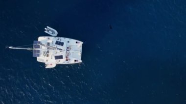 Hava görüntüsü, nefes kesen bir deniz manzarasının ortasında katamaran yelkenliyi yakalıyor. Güneş panelleri ile donatılmış el değmemiş beyaz bir gemi ve küçük şişme bir sandal sergiliyor.