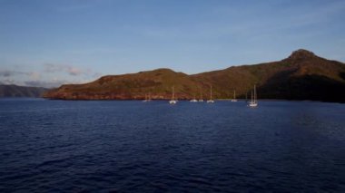 İnsansız hava aracı görüntüleri sakin, derin mavi bir denize demir atmış yelkenlilerin sakin bir sahnesini yakalıyor. Yavaş yavaş uçup giderken ön planda geniş bir su birikintisi ve güneşli bir tepe ortaya çıkıyor.