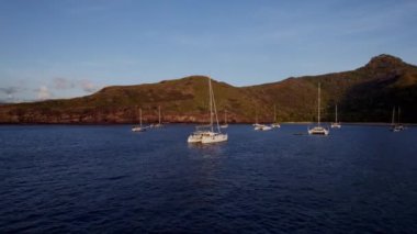 Bir hava manzarası sakin, derin mavi bir deniz üzerinde yüzen yelkenlileri yakalar, arka planda tepe yeşil bir manzara, yumuşak güneş ışığı ise sakin sahil sahnesinde sıcak bir parıltı oluşturur.