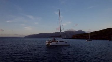 Sakin suların üzerinde süzülen sakin bir yelkenli manzarası, yumuşak alacakaranlıkta yuvarlanan tepeler tarafından çerçevelenmiş, mavi ve gri tonlarda