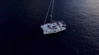 Resimli bir sahne, yükselen bir dron perspektifinden sakin bir koyda demirlemiş yelkenlileri yakalıyor, ön planda yelkenleri tamamen açılmış beyaz bir yelkenliyi ön plana çıkarıyor.