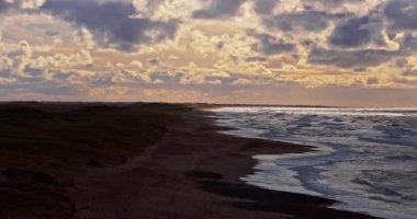 Vesterhavet 'in güneşli sahil şeridinden, okyanus dalgalarının ritmik dansına pürüzsüz bir şekilde dönüşerek kıyıya doğru süzülen sağa doğru bir bakış açısıdır.