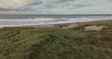 Vesterhavet 'in kıyı şeridine tepeden bakan yeşil çimlerle kaplı kum tepeleri. Köpüklü dalgalar nemli kumlara dökülüyor.