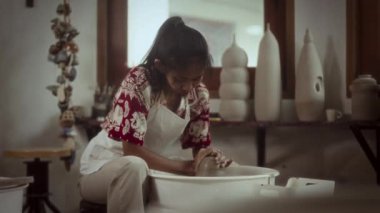 Çömlekçinin çarkında kil şekillendiren odaklanmış bir kadın dikkatle çömlek parçasını özenle hazırlıyor.