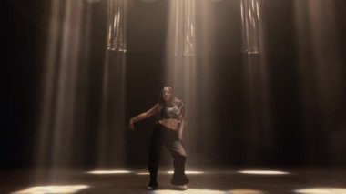 Bir kızın hareketli, ritmik hareketleri yoğun ışın ışıklarıyla keskin bir şekilde aydınlatılır ve karanlık odada dramatik bir görüntü yaratır.