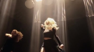 Düşük açılı POV 'tan, iki kız karanlık bir stüdyoda ışık huzmeleri altında çağdaş bir dans gerçekleştiriyor.